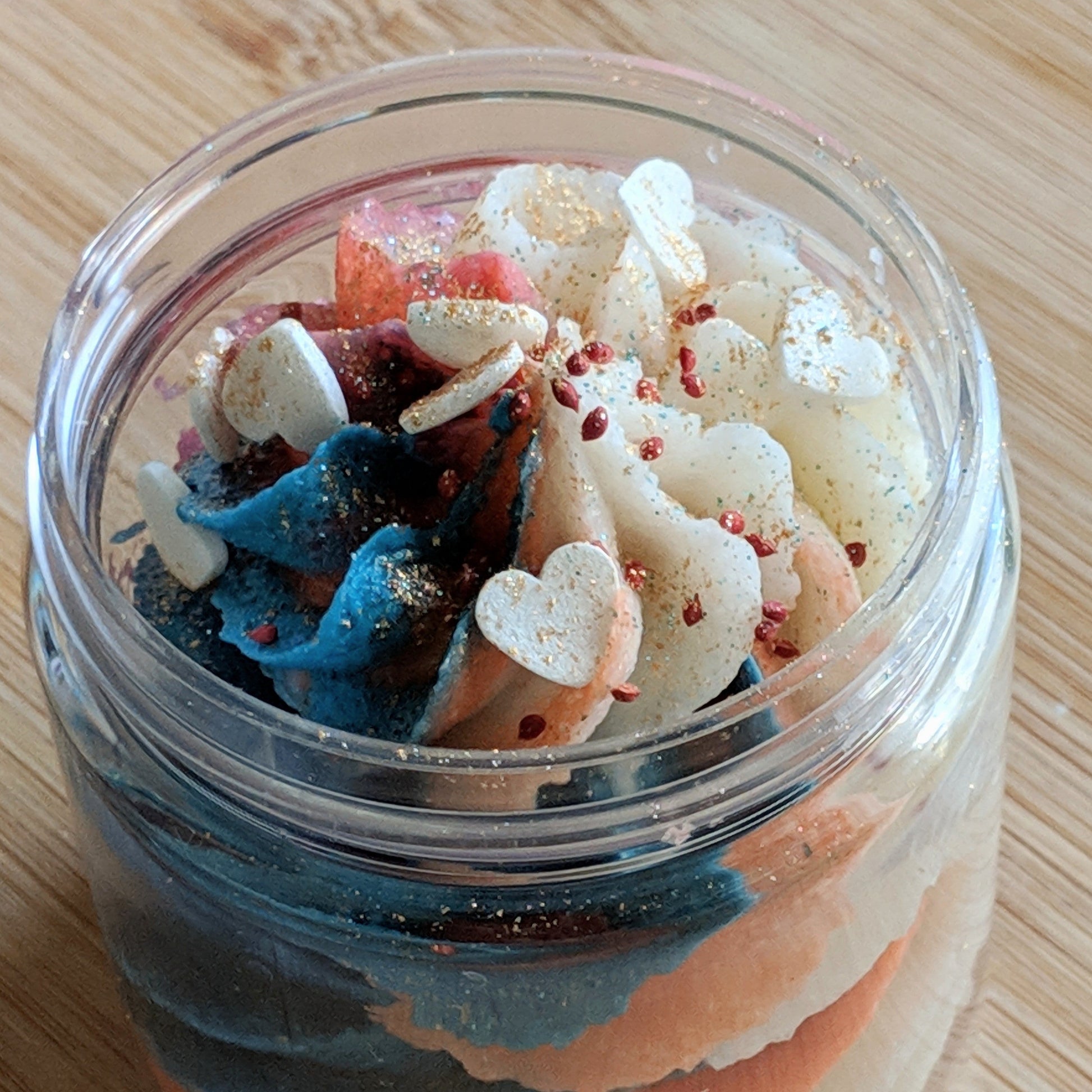 A jar of Peach Blossom Whipped Sugar Scrub by PI Colors sits on a wooden surface, showcasing its swirled whipped cream in blue, pink, and white hues. It is adorned with white heart-shaped sprinkles and glitter, offering a luxurious bath experience with its organic cane sugar formulation.