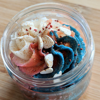 A jar adorned with red, white, and blue swirls of frosting, topped with almond flakes and red sprinkles, sits on a wooden surface, much like the exquisite Peach Blossom Whipped Sugar Scrub by PI Colors. Enriched with organic cane sugar, it offers a luxurious sensory experience.