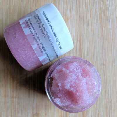 An open jar of PI Colors Lavender Lemonade Sugar Lip Scrub sits on a wooden surface, showing its grainy texture, while another nearby jar displays the organic label and ingredients.