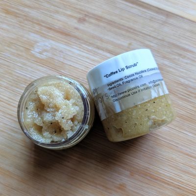 On a wooden surface, there are two small jars; one is open, while the other is closed. The open jar displays a gritty, beige scrub intended for exfoliating lips with organic ingredients. The closed jar bears the label "Coffee Sugar Lip Scrub" from PI Colors and highlights its rich, aromatic coffee-flavored components designed to treat your lips.