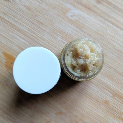 An open small glass jar containing PI Colors' Coffee Sugar Lip Scrub, a grainy, beige substance with a coarse texture, rests on a wooden surface next to its white lid. This organic lip scrub is crafted to gently and effectively exfoliate lips with a delightful coffee flavor.