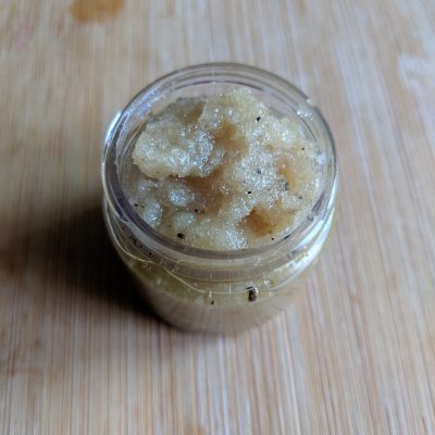 A compact glass jar of PI Colors Coffee Sugar Lip Scrub rests on a wooden surface. The organic ingredients give it a grainy texture with dark specks scattered throughout. With the lid removed, its exfoliating contents, crafted to rejuvenate your lips, are on full display.