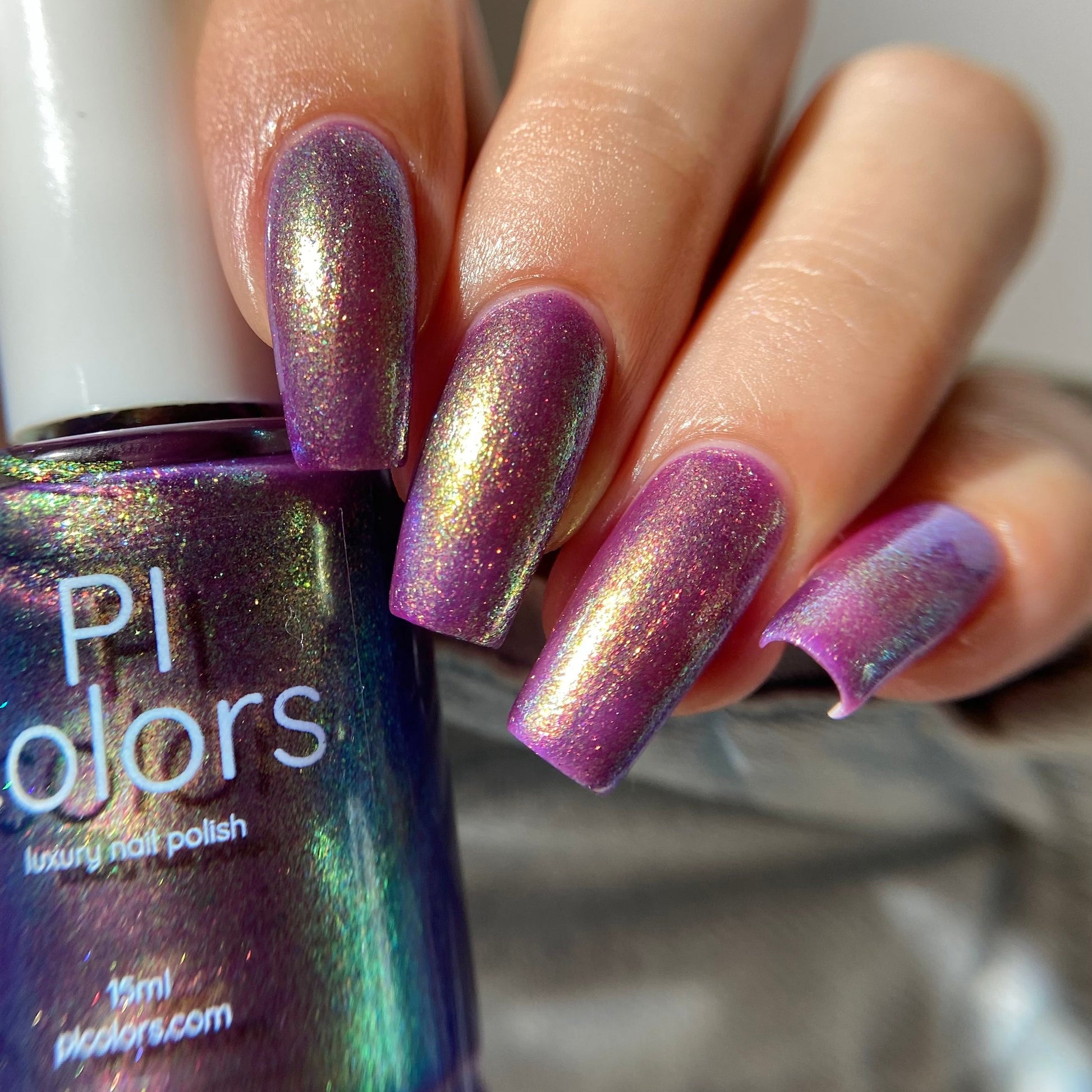 A close-up of a hand with manicured nails painted in a shimmering iridescent purple nail polish, holding a bottle of Pi Colors luxury nail polish.