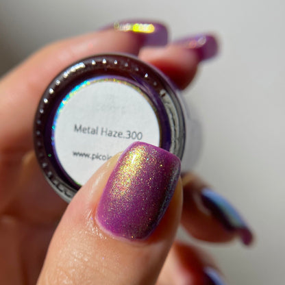 A close-up of a hand holding a small jar of nail polish. The nail polish being demonstrated is a metallic purple with a shimmering gold finish, labeled 'Metal Haze.