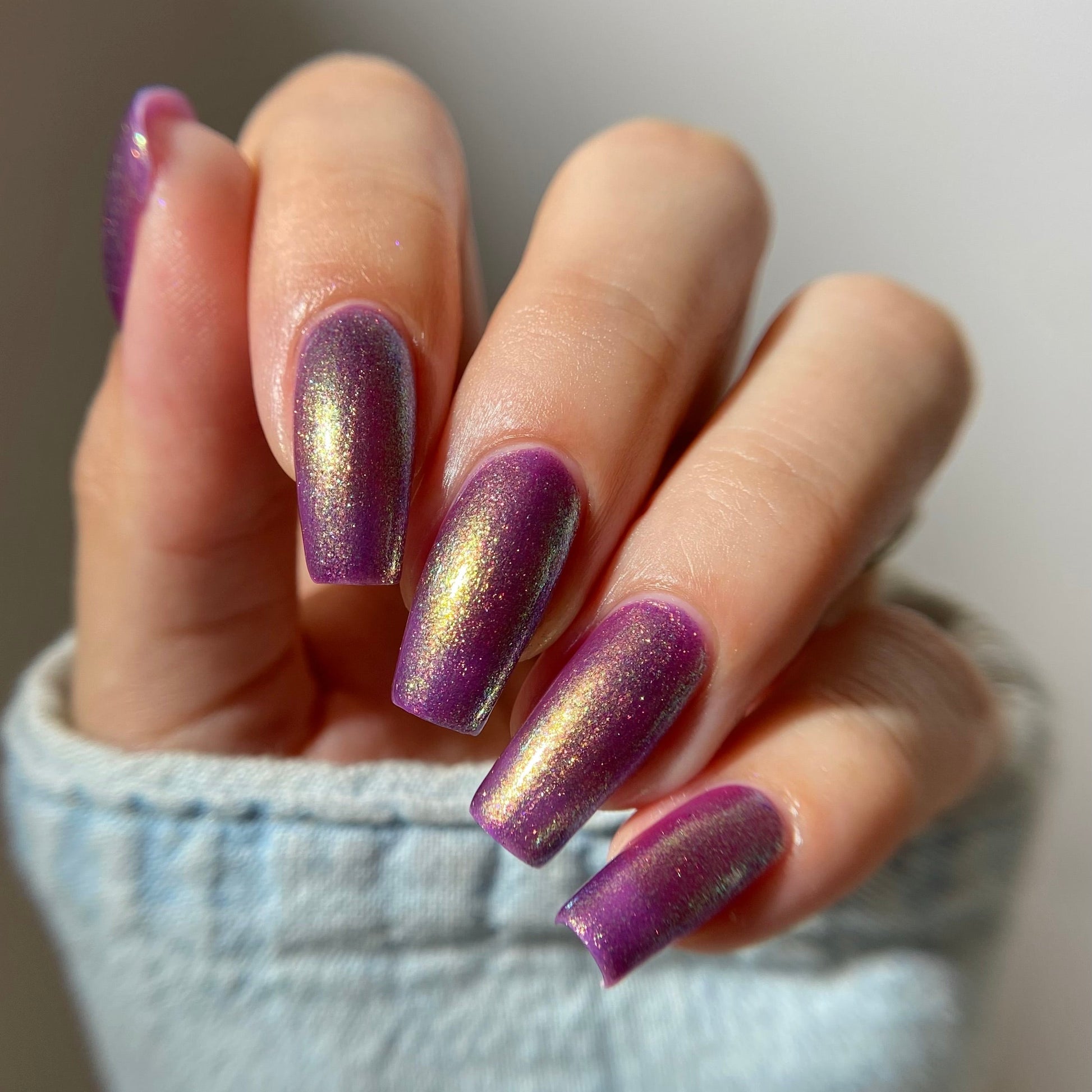 A close-up of a hand with manicured nails painted in a metallic purple polish with a shimmering gold foil effect, resting on a light blue denim sleeve.
