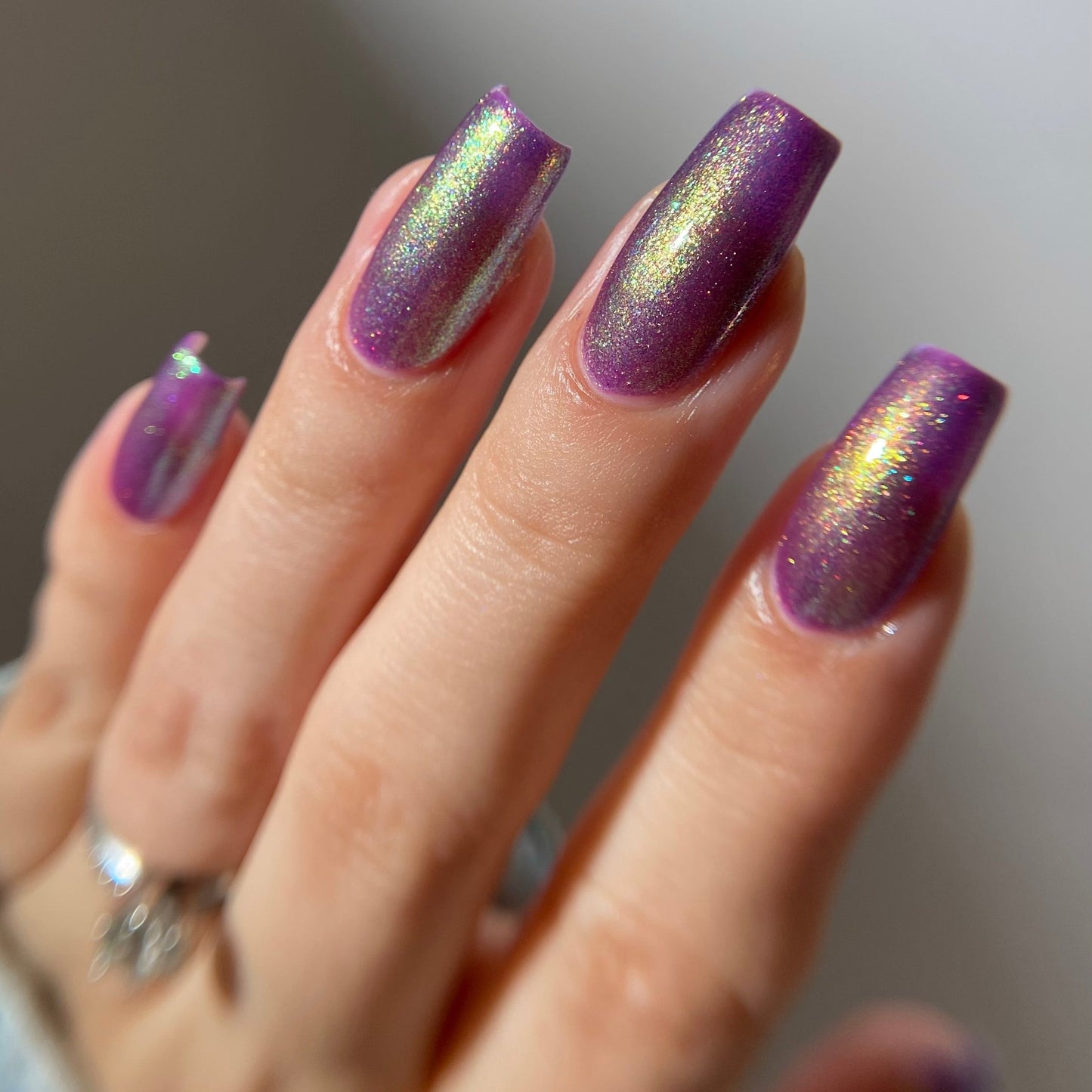 A close-up of a hand with long, coffin-shaped nails painted in a shimmering metallic purple nail polish with an iridescent holographic finish reflecting gold and green tones.