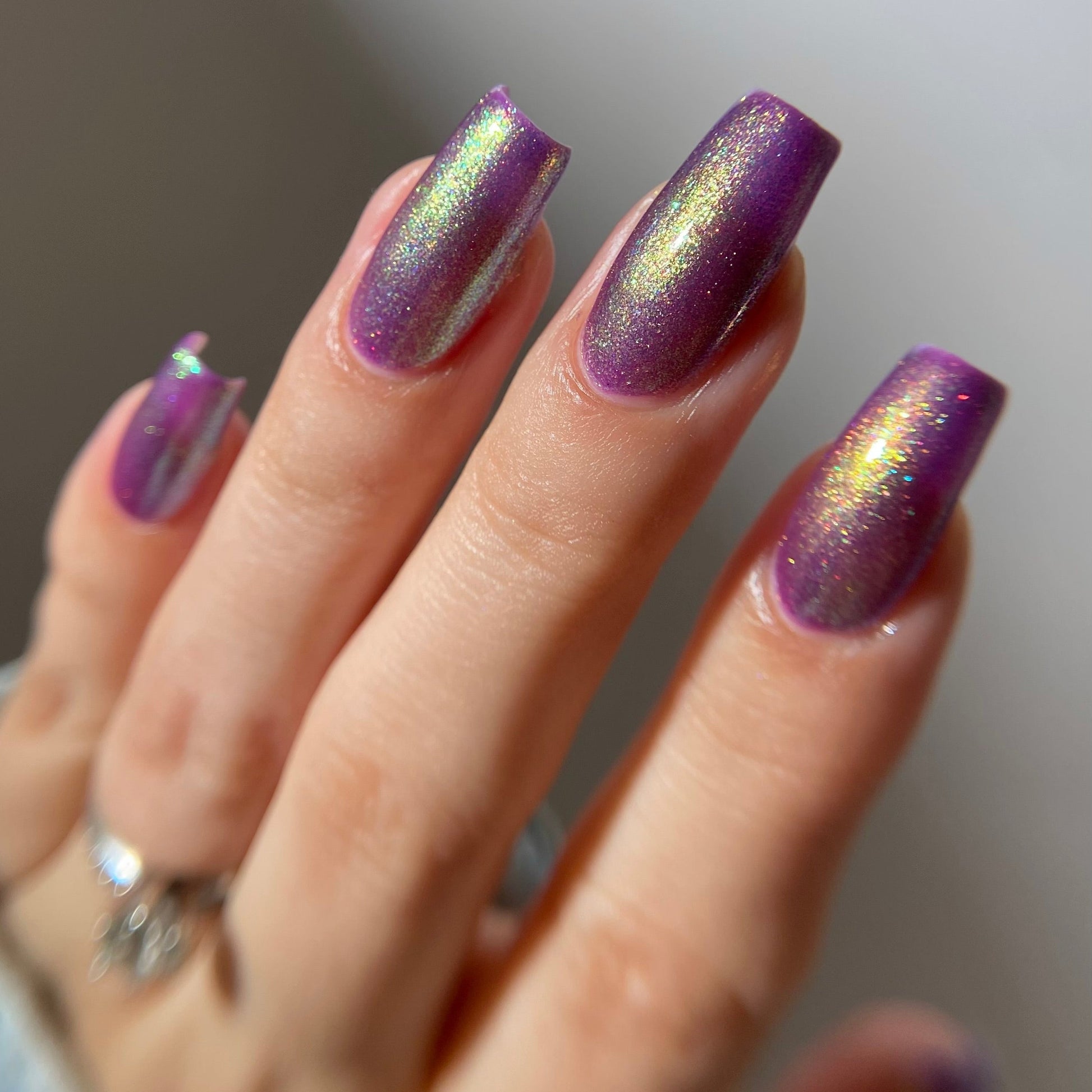 A close-up of a hand with long, coffin-shaped nails painted in a shimmering metallic purple nail polish with an iridescent holographic finish reflecting gold and green tones.