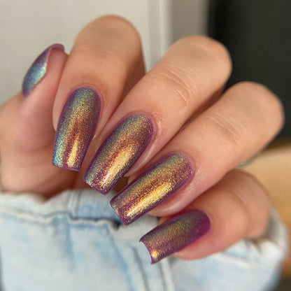 A close-up of a hand with almond-shaped nails painted in a shimmering holographic nail polish that shifts between purple, gold, and blue-green, wearing a light blue denim jacket.