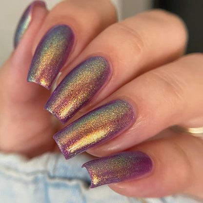 A close-up of a hand with manicured nails painted in a shimmering iridescent nail polish that shifts between purple, gold, and green, resting against light denim fabric.