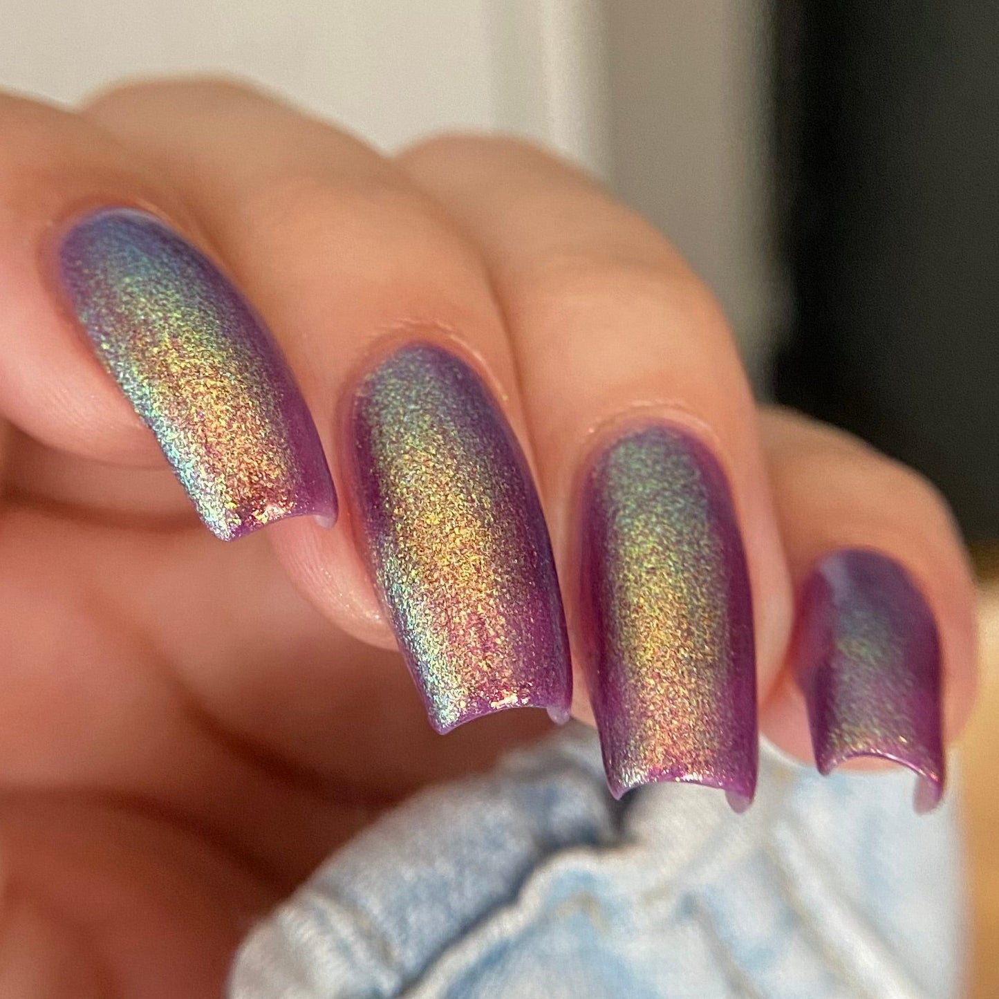 A close-up of a hand with almond-shaped nails painted in a holographic iridescent nail polish that shifts between purple, blue, green, and gold. The hand is resting near light-wash denim jeans.