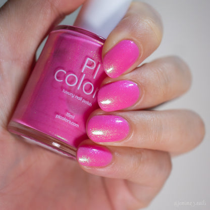 A hand holds a bottle of PI Colors Mint and Summer.100 handmade nail polish, with nails painted in vibrant pink and topped with a shimmery, glittery finish to match.