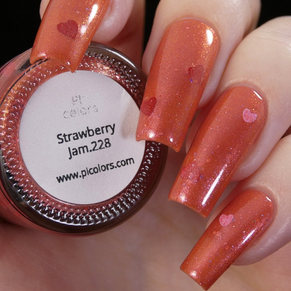 Close-up of a hand with long, glossy nails in PI Colors' Strawberry Jam.228 red, adorned with small heart designs. The luxury nail lacquer bottle, labeled "Strawberry Jam.228," rests between the fingers.