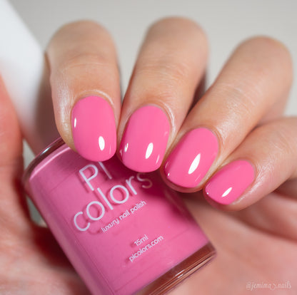 A hand with neatly manicured nails painted in glossy pink Neapolitan.432 by PI Colors holds a matching bottle of this luxury nail lacquer.
