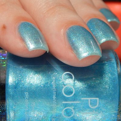 The image captures a person's fingers adorned with Loulaki.101 Blue Nail Polish from PI Colors, featuring a shimmering metallic blue hue with iridescent flakes. In the foreground, there's a partial view of the nail polish bottle, expertly complementing the stunning nails.