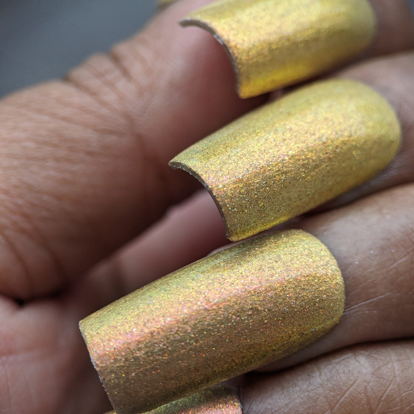 Close-up of a hand showcasing PI Colors' Golden Fuyuki.000 luxury nail polish, featuring long, shimmering nails with a metallic gold holo glitter finish, set against a blurred background. The well-manicured nails catch the light, highlighting their sparkle and exquisite golden multichrome allure.