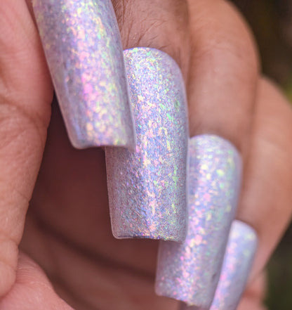 A close-up reveals a hand with long nails painted in PI Colors' Iris Sky.005, featuring lilac purple hues and iridescent rainbow flakies that shimmer in pink and blue. The textured nails glisten against a softly blurred background.