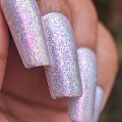 Close-up of a hand with long, squared acrylic nails featuring PI Colors Iris Sky.005 nail polish, a lilac purple shade with rainbow flakies creating an iridescent holographic effect in pink and blue against a blurred backdrop.