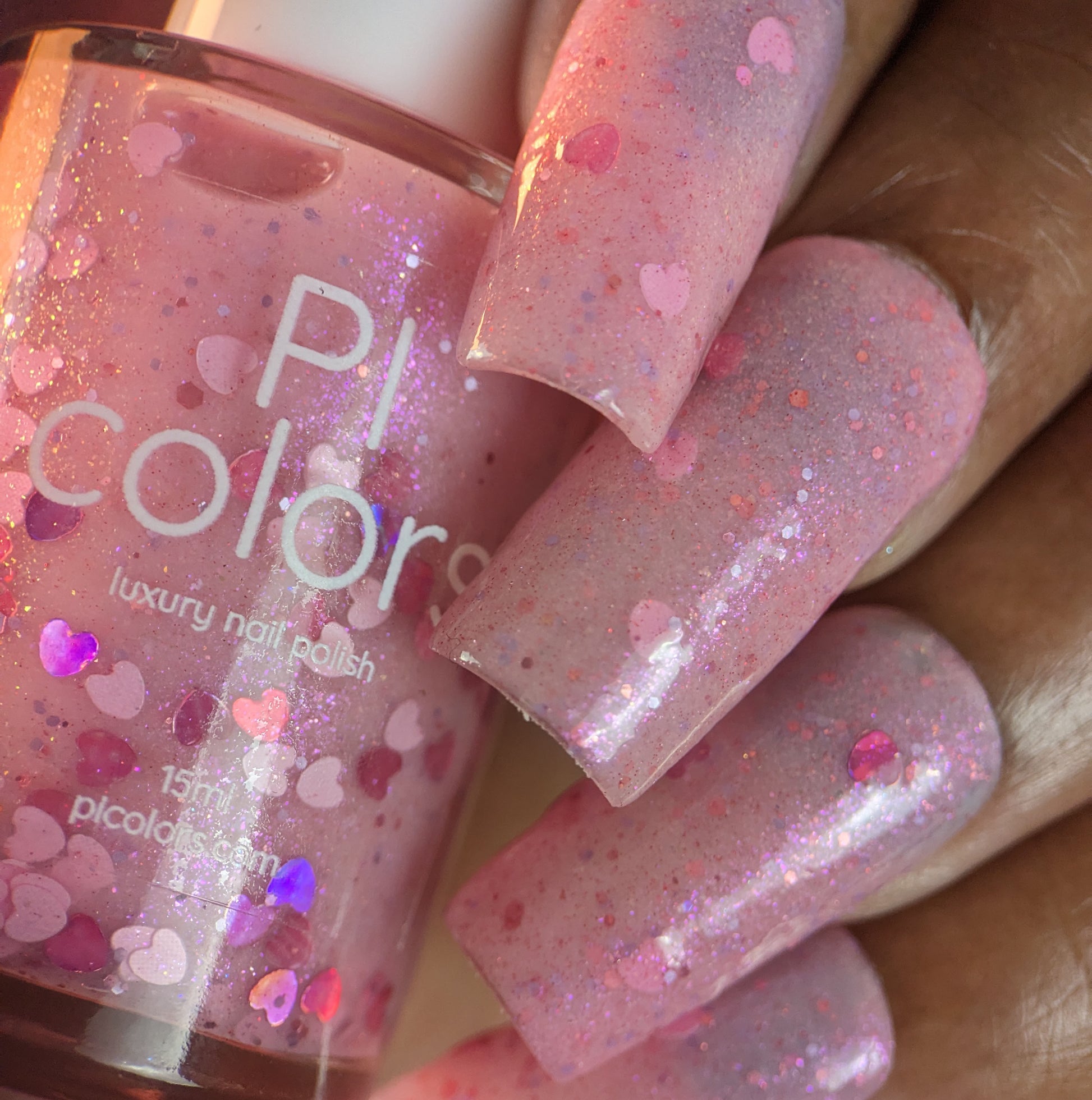 A close-up features a hand with long nails painted in pale pink "Pink Berry Heart.200" polish by PI Colors, adorned with holographic hearts. Beside the dazzling nails sits the bottle proudly displaying its 10-free formula.