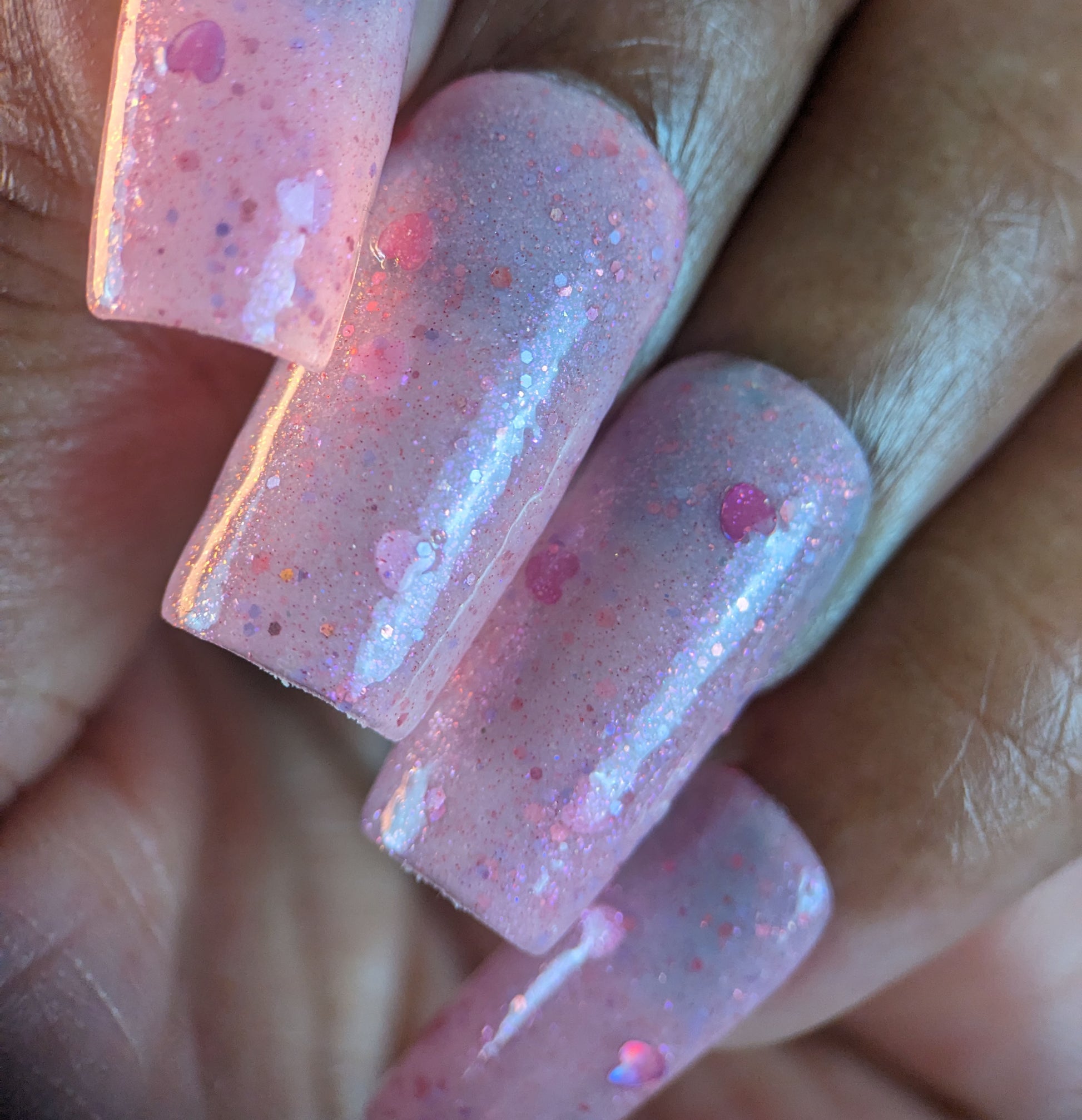 Close-up of fingers with long, square-shaped nails painted in PI Colors’ Pink Berry Heart.200, showcasing sparkly glitter and holographic hearts. The glossy finish enhances circular pink accents against a blurred background, providing a dazzling 10-free manicure style.