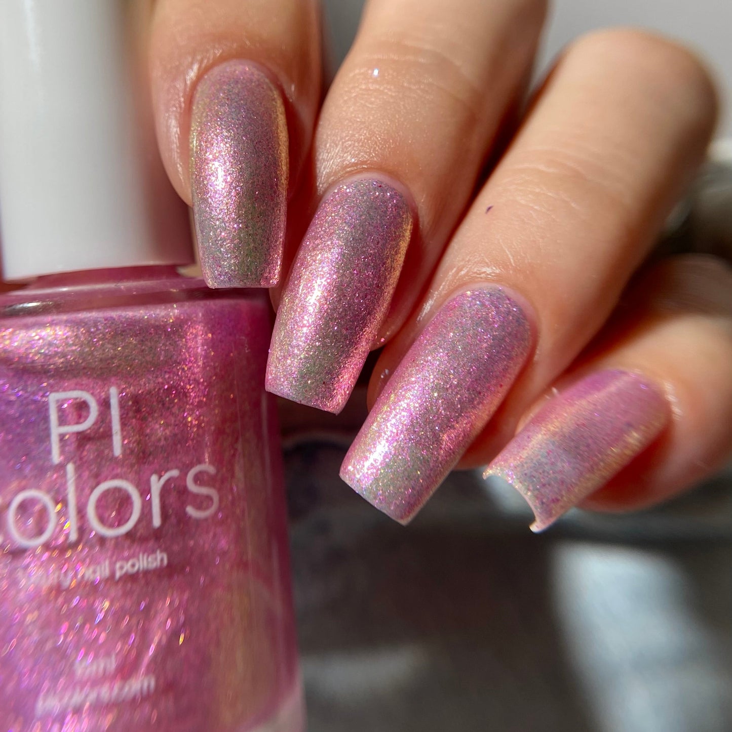 A close-up of a hand with fingernails painted in a shimmering pink nail polish, holding a bottle of PI colors nail polish with the same glittery pink shade.