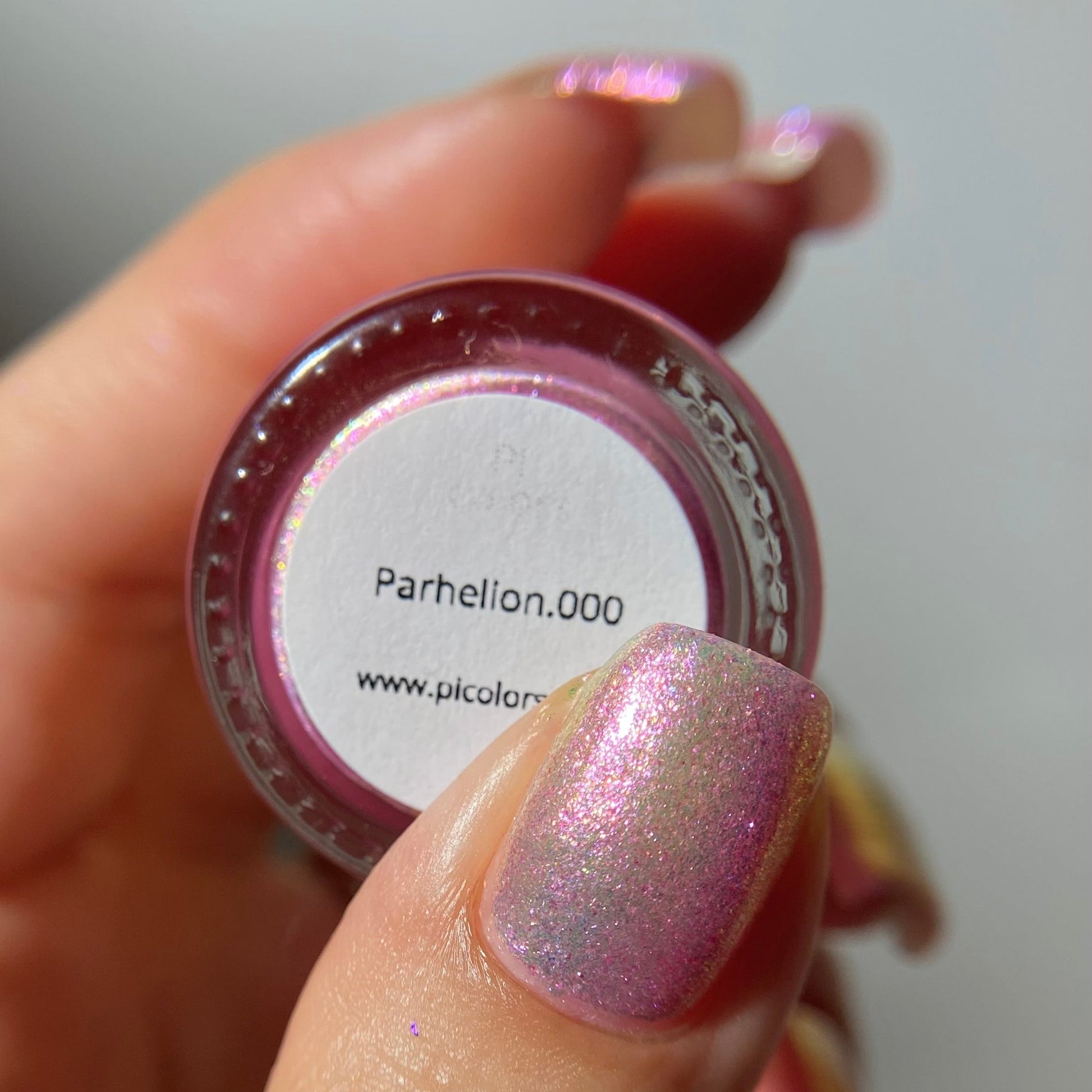 A close-up of a hand holding a small round pot of shimmering pink nail polish or pigment. The label reads 'Parhellen.