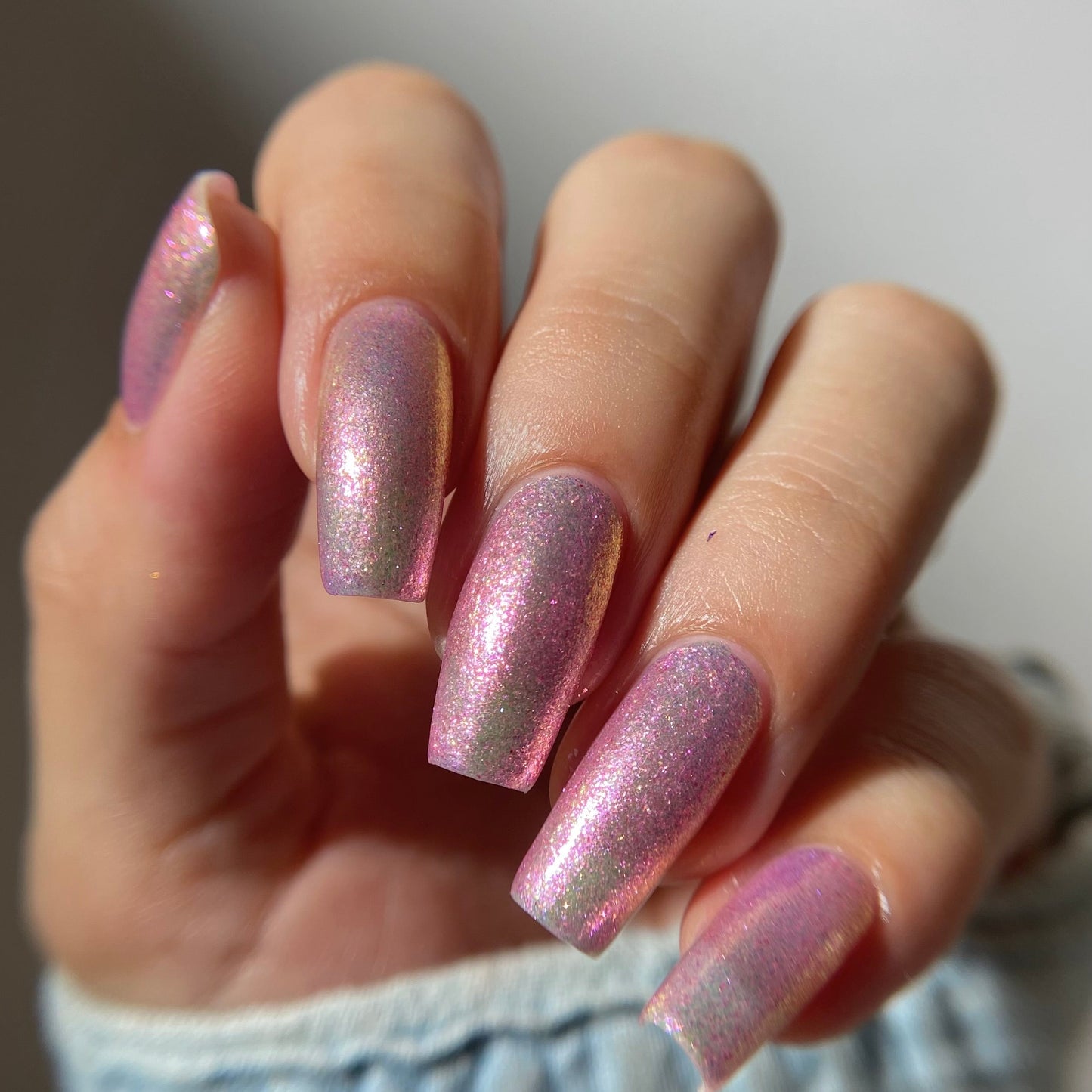 A close-up of a hand with long, almond-shaped nails painted in a shimmering pink nail polish with a subtle iridescent gold shimmer, wearing a light blue denim sleeve.