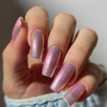 A close-up of a hand with long, almond-shaped nails painted in a shimmering pink nail polish with a subtle iridescent gold shimmer, wearing a light blue denim sleeve.