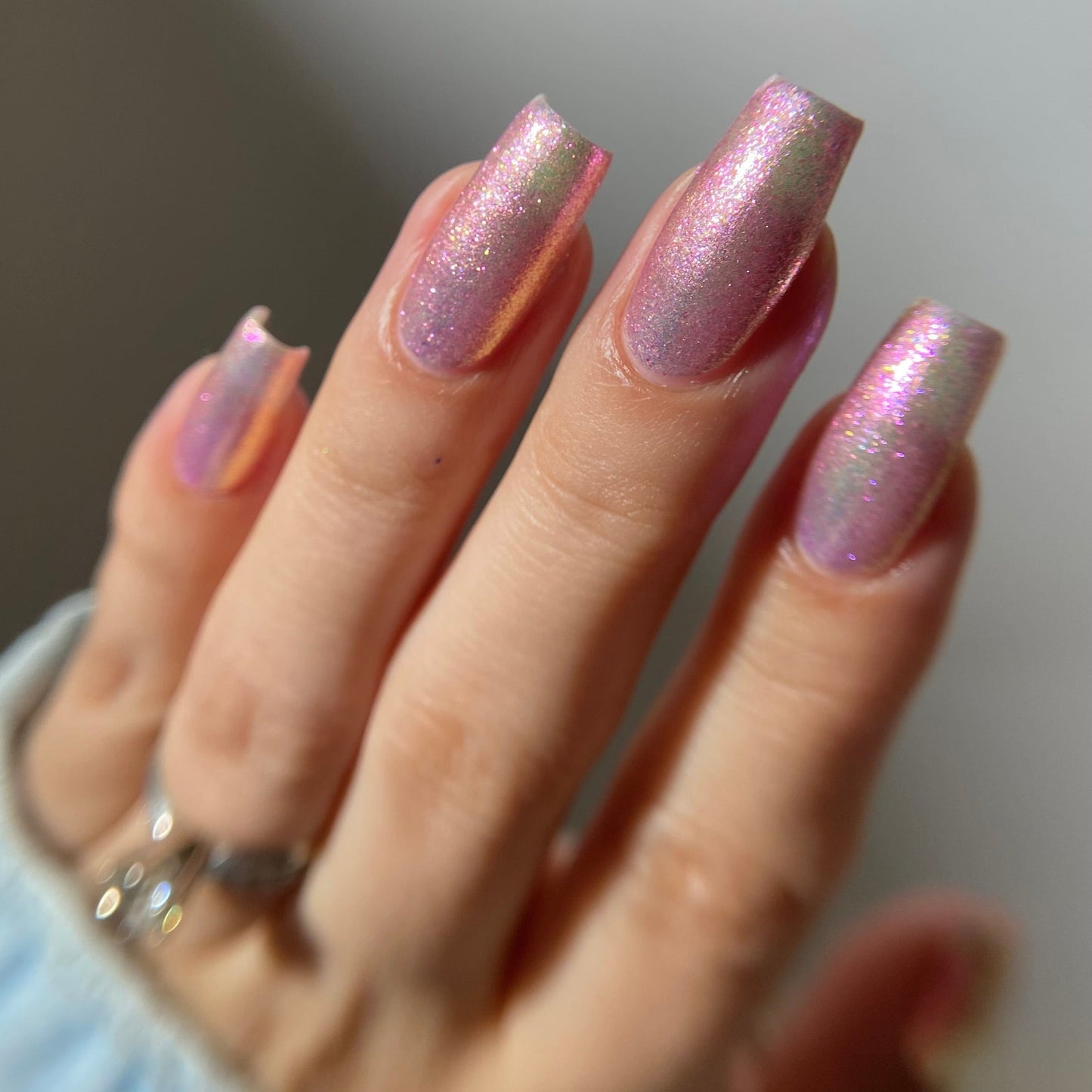 A close-up of a hand with manicured nails painted in a shimmering holographic pink polish, wearing a silver ring on one finger.