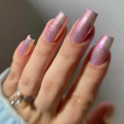 A close-up of a hand with manicured nails painted in a shimmering holographic pink polish, wearing a silver ring on one finger.