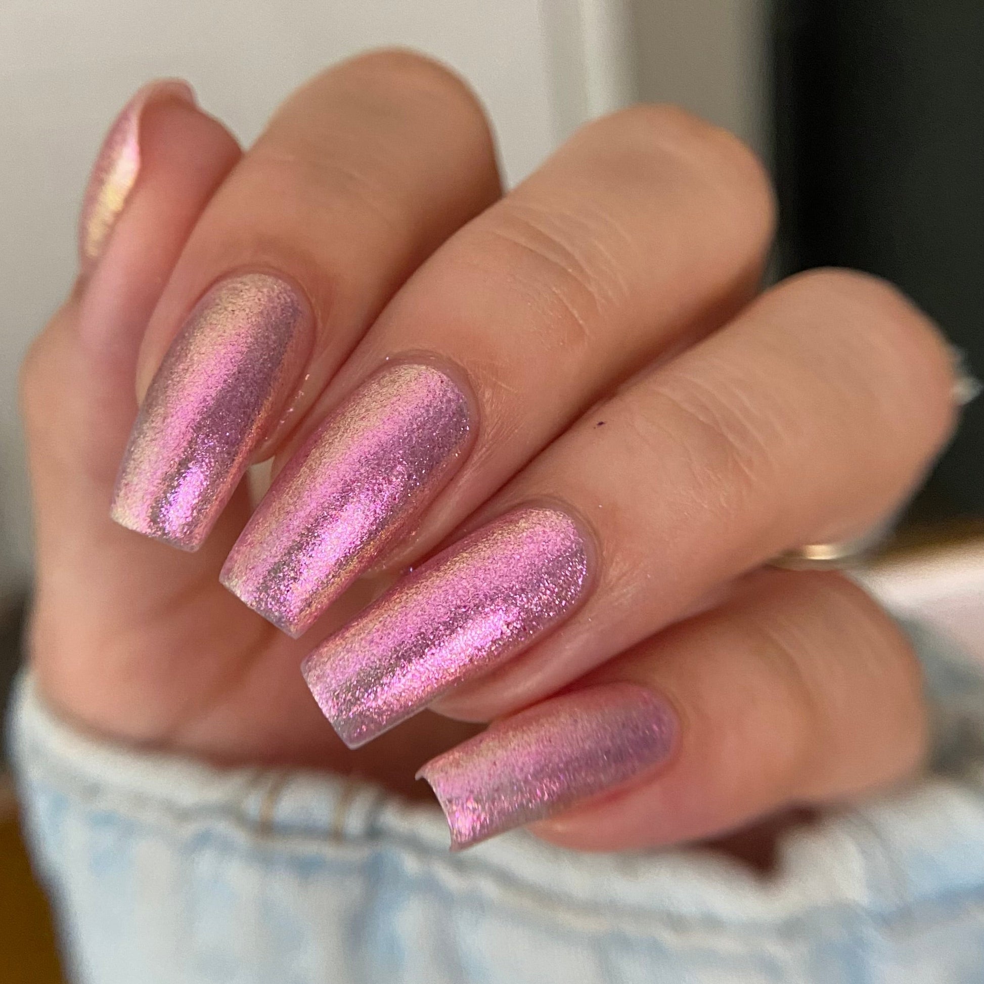 A close-up of a hand with long, square-shaped nails painted in a shimmering metallic pink nail polish with a holographic sheen.