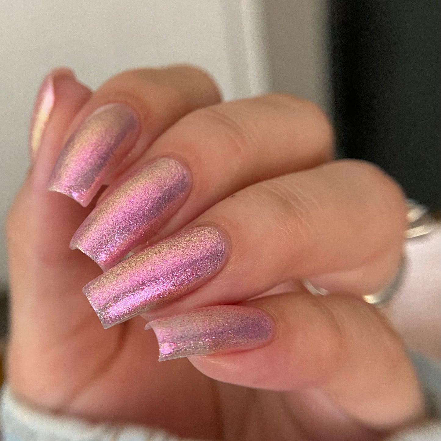 A close-up of a hand with long, square-shaped nails painted in a shimmering metallic pink nail polish with a holographic iridescent finish.