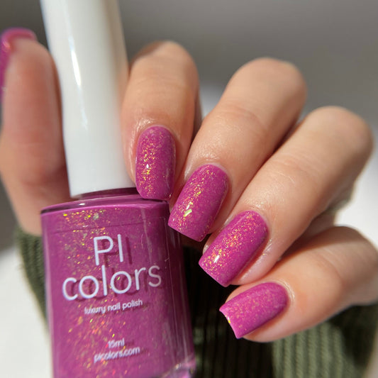 A hand with nails painted in sparkly pink holds a "PI Colors" bottle of "Phoenician Dusk.000" handmade nail polish. The magenta and purple base gleams on neatly manicured nails against a softly blurred backdrop, highlighting its 10-free formula allure.