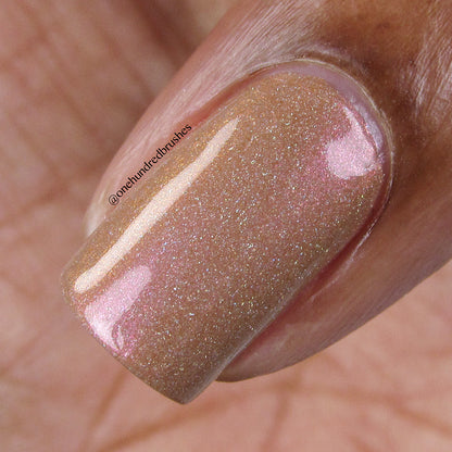 A close-up image showcases a fingernail adorned with PI Colors' Phoenix.087 Brown Nail Polish, a shimmering gold and pinkish duochrome shade. This 10-free polish offers a glossy finish complemented by a subtle holographic effect, with the skin around the nail also visible.