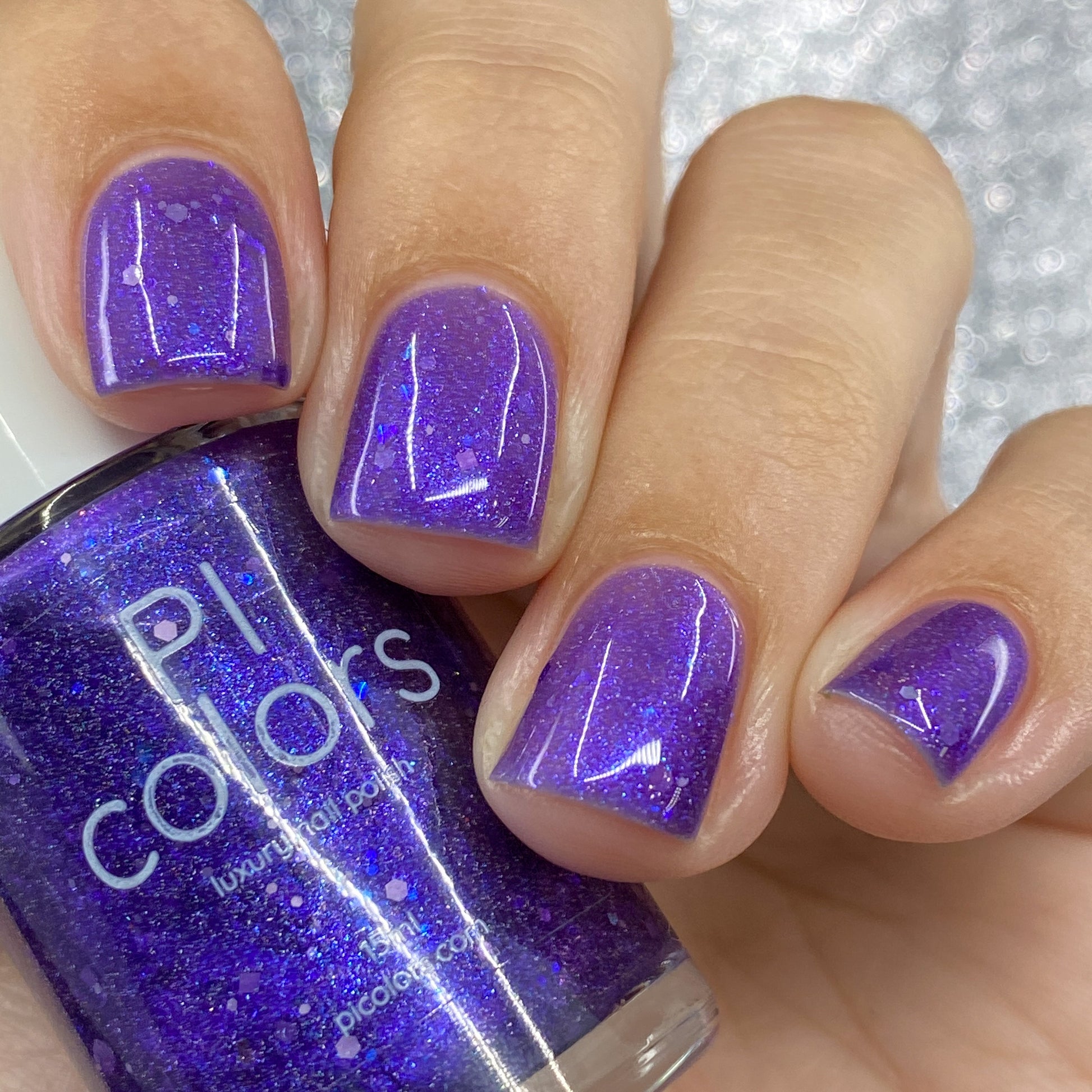 A hand with short, well-manicured nails flaunts a vibrant purple nail polish adorned with dazzling holo glitters. Nearby, a bottle of this same polish features the label "PI Colors" with the product name "Purple Smoke.001 Purple Nail Polish." The background shimmers with a sparkling, out-of-focus glitter texture.