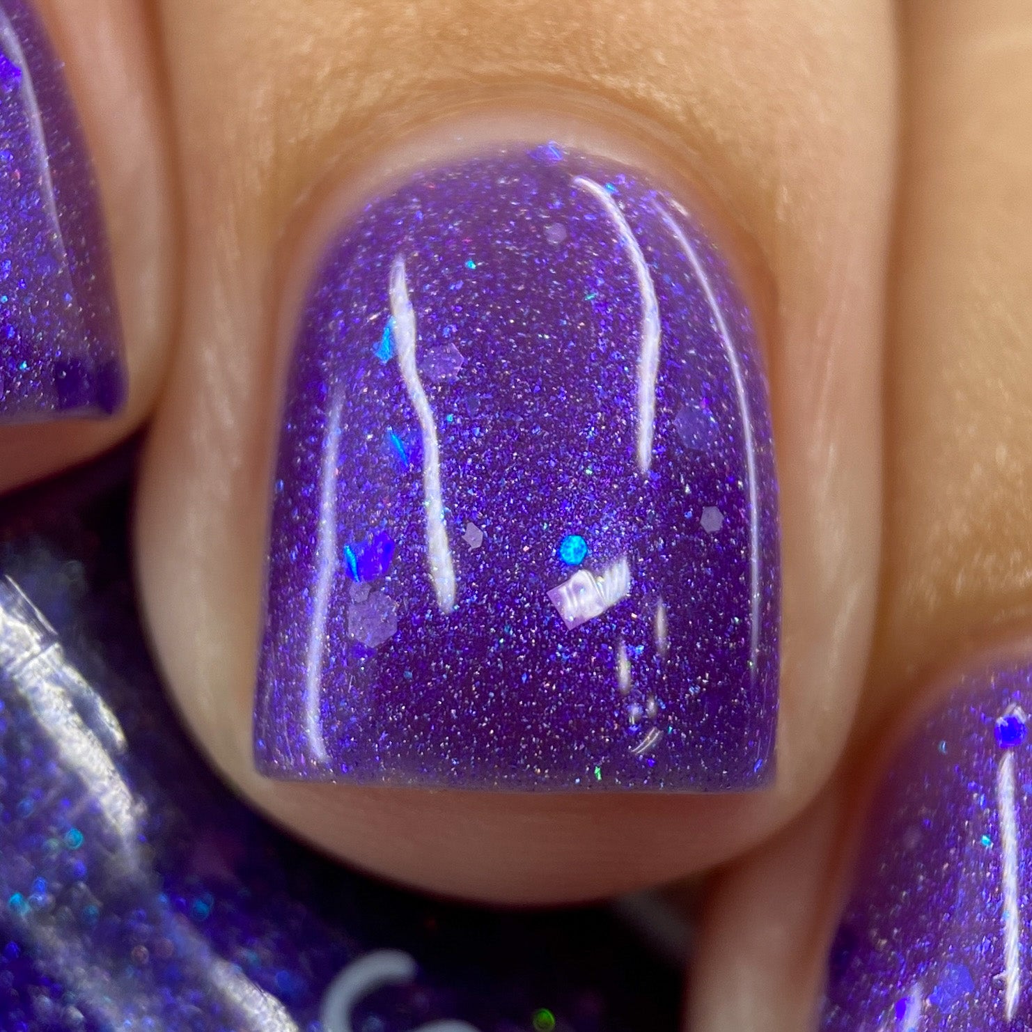 A close-up of a fingernail showcases PI Colors' Purple Smoke.001 Purple Nail Polish, featuring an iridescent purple jelly finish embellished with holographic purple glitters for a metallic sheen. The nail is flawlessly manicured, with hints of two additional nails visible on either side.