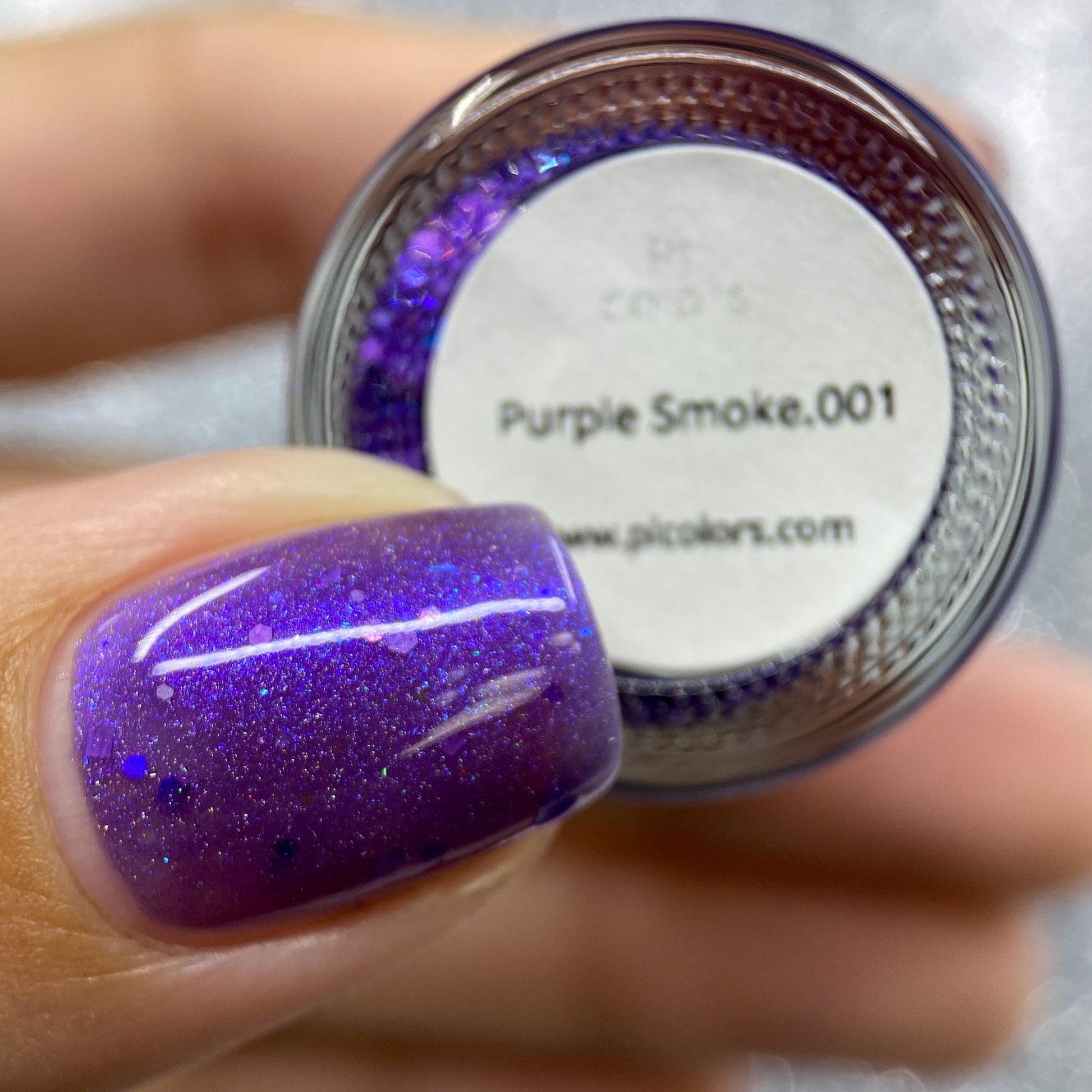 Close-up of a fingernail adorned with sparkly holo purple glitters, held next to a small container labeled "Purple Smoke.001 Purple Nail Polish" from PI Colors. The glossy nail polish gleams with glittery specks against the brand's website displayed below.