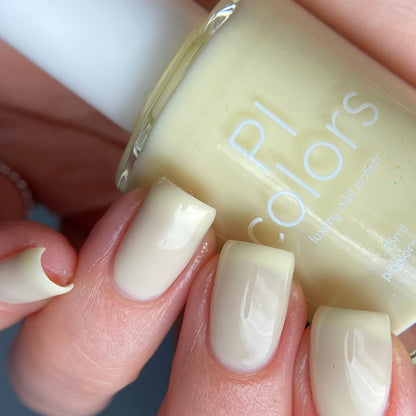 A close-up of a person's hand with neatly manicured nails painted in pastel yellow captures the essence of elegance. The hand holds a bottle of Chiffon.400 Yellow Nail Polish by PI Colors, showcasing its luxurious creme finish. A blurred background emphasizes the vibrant nails and polish bottle.