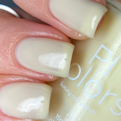 Close-up of a hand with nails painted in a glossy, pale cream color. The PI Colors bottle of Chiffon.400 Yellow Nail Polish is partially visible, showcasing the words "luxury nail polish." The creme finish offers a smooth and shiny appearance.