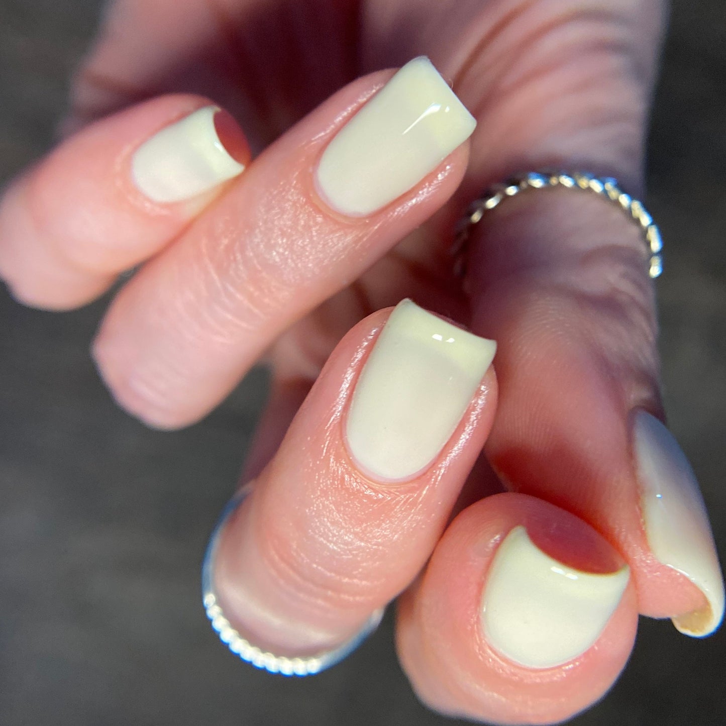 Close-up of a hand featuring squared-off, glossy nails painted in pastel yellow using PI Colors' Chiffon.400 Yellow Nail Polish. The fingers are adorned with delicate silver rings with a chain-like design, and the softly blurred background accentuates the vibrant nail art and shimmering jewelry.