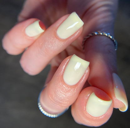 Close-up of a hand showcasing PI Colors' Chiffon.400 Yellow Nail Polish from the Spring Collection, with neatly manicured pale yellow nails. The fingers are adorned with simple silver rings against a softly blurred background, highlighting the glossy finish of this 10-free nail polish.