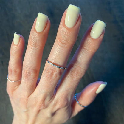 A hand with manicured nails painted in Chiffon.400 Yellow Nail Polish by PI Colors is shown. The fingers, adorned with delicate silver rings including a thin band with small gems, stand out against a soft, blurred dark surface.