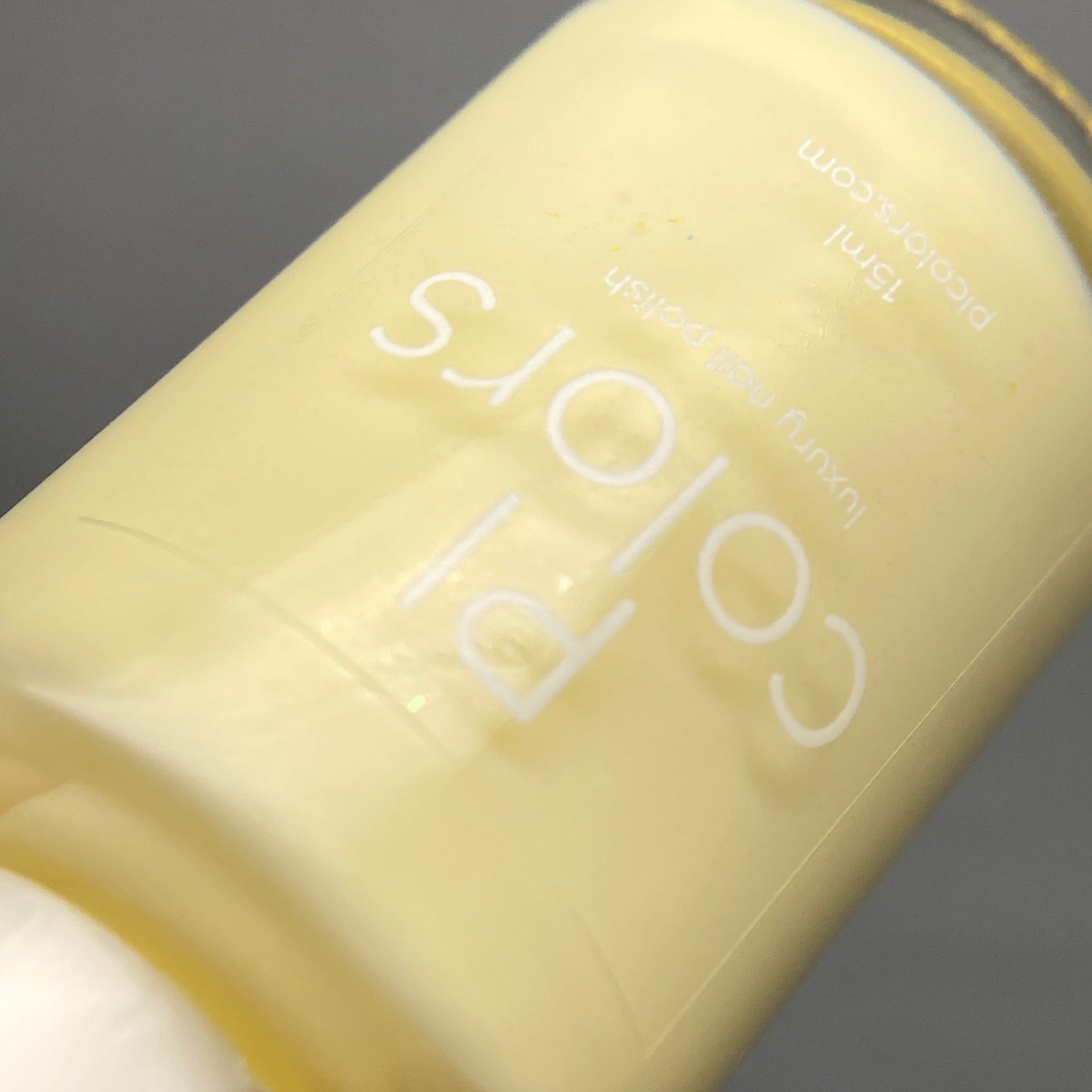 A close-up shot of a 15 ml bottle of Chiffon.400 Yellow Nail Polish by PI Colors, featuring its luxurious pastel yellow hue and crème finish, is artistically tilted sideways against a gray background.