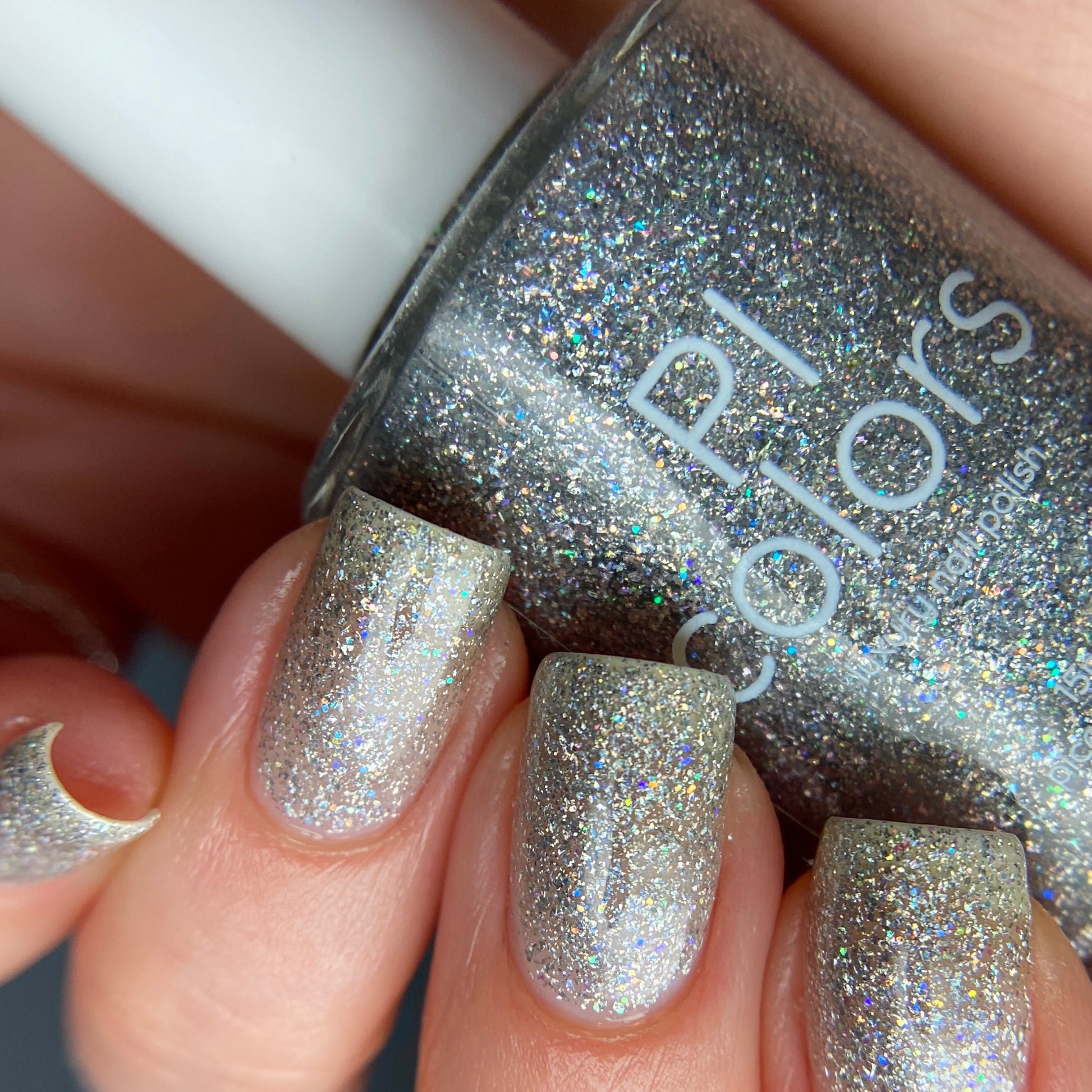 Close-up of a hand showcasing nails adorned with luxury nail lacquers in the shimmering "Diamond Blast.401 Silver Holographic Glitter" by PI Colors. In the background, a bottle of this 10-free nail polish topper can be seen, enhancing the scene with its glittery allure.