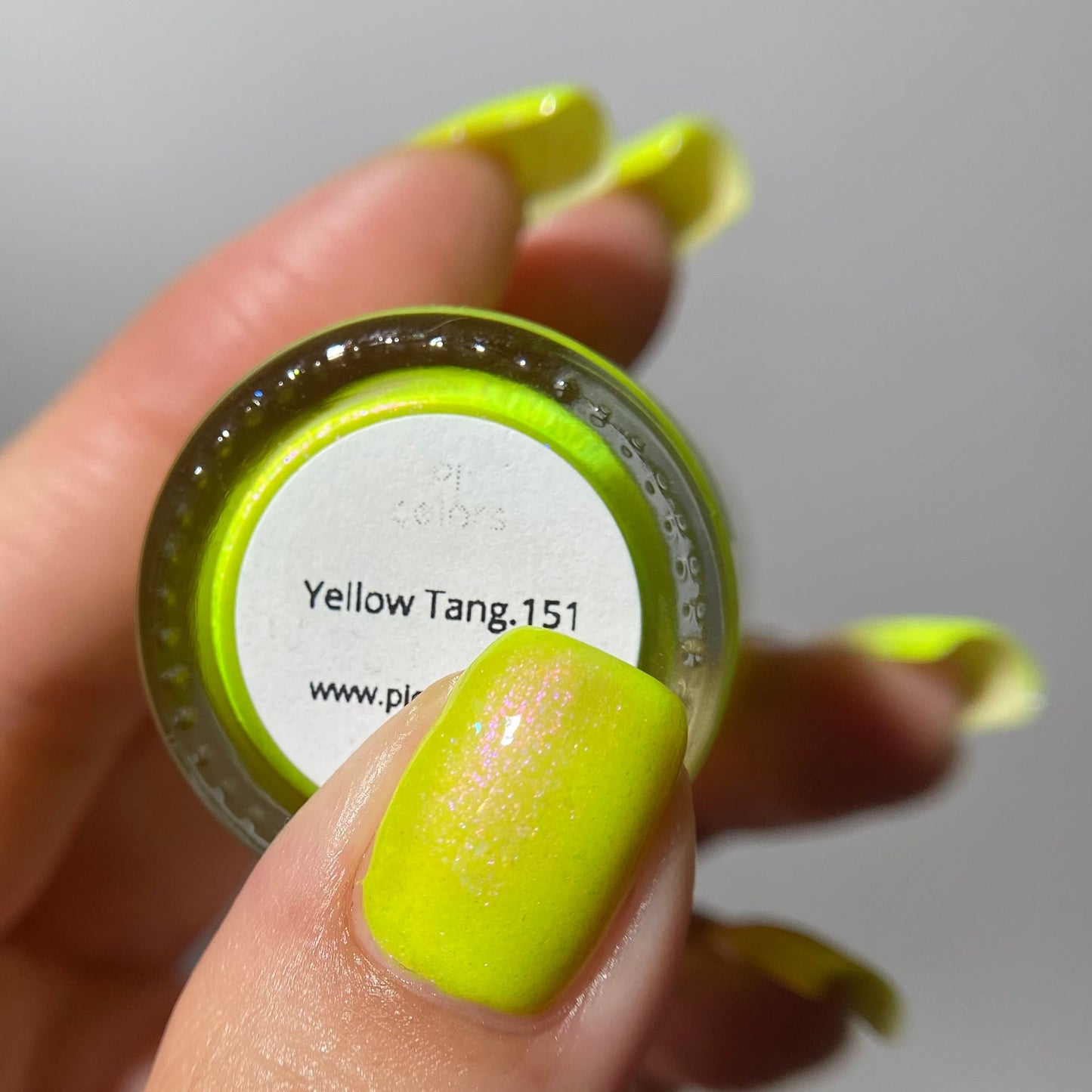 A close-up of a hand holding a small round jar of nail polish or pigment. The product color is a bright neon yellow-green, matching the swatch labeled 'Yellow Tang.