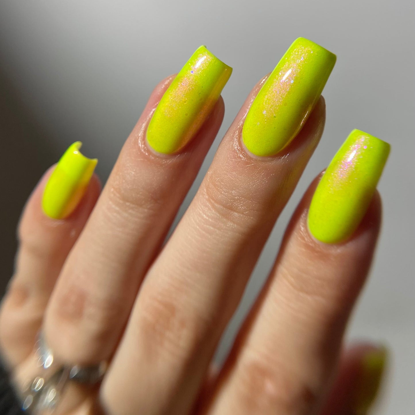 A close-up of a hand with long, square-shaped nails painted in a bright neon yellow-green polish with subtle pink glitter flecks, wearing silver rings against a soft gray background.