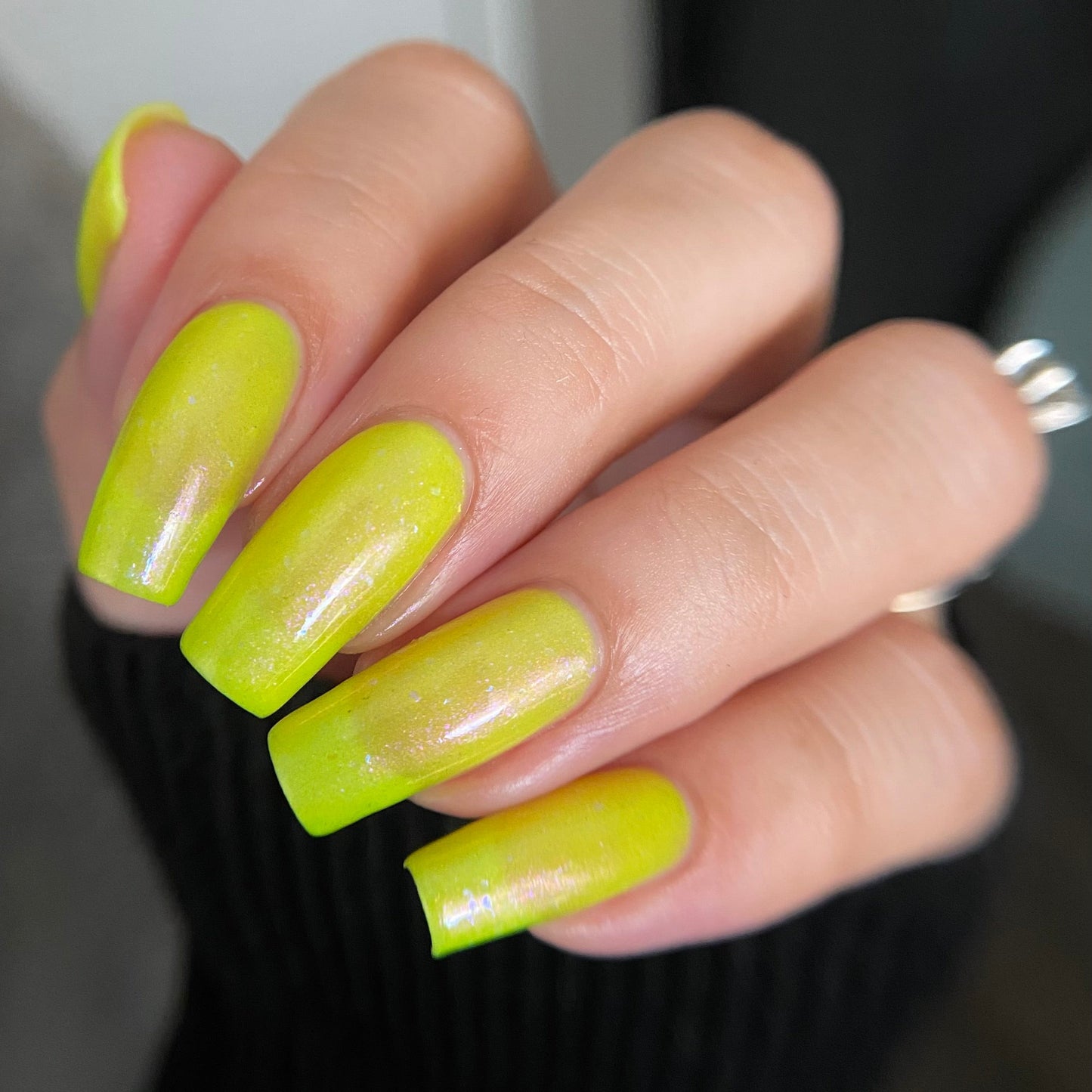A close-up of a hand with long, glossy, bright neon yellow manicured nails, wearing a silver ring, set against a dark ribbed sweater and a soft gray background.