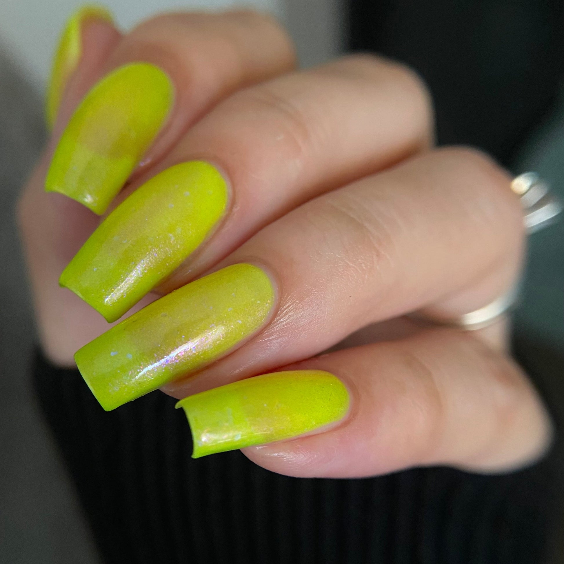 A close-up of a hand with long, glossy, bright neon yellow manicured nails, wearing a silver ring against a dark ribbed sweater.