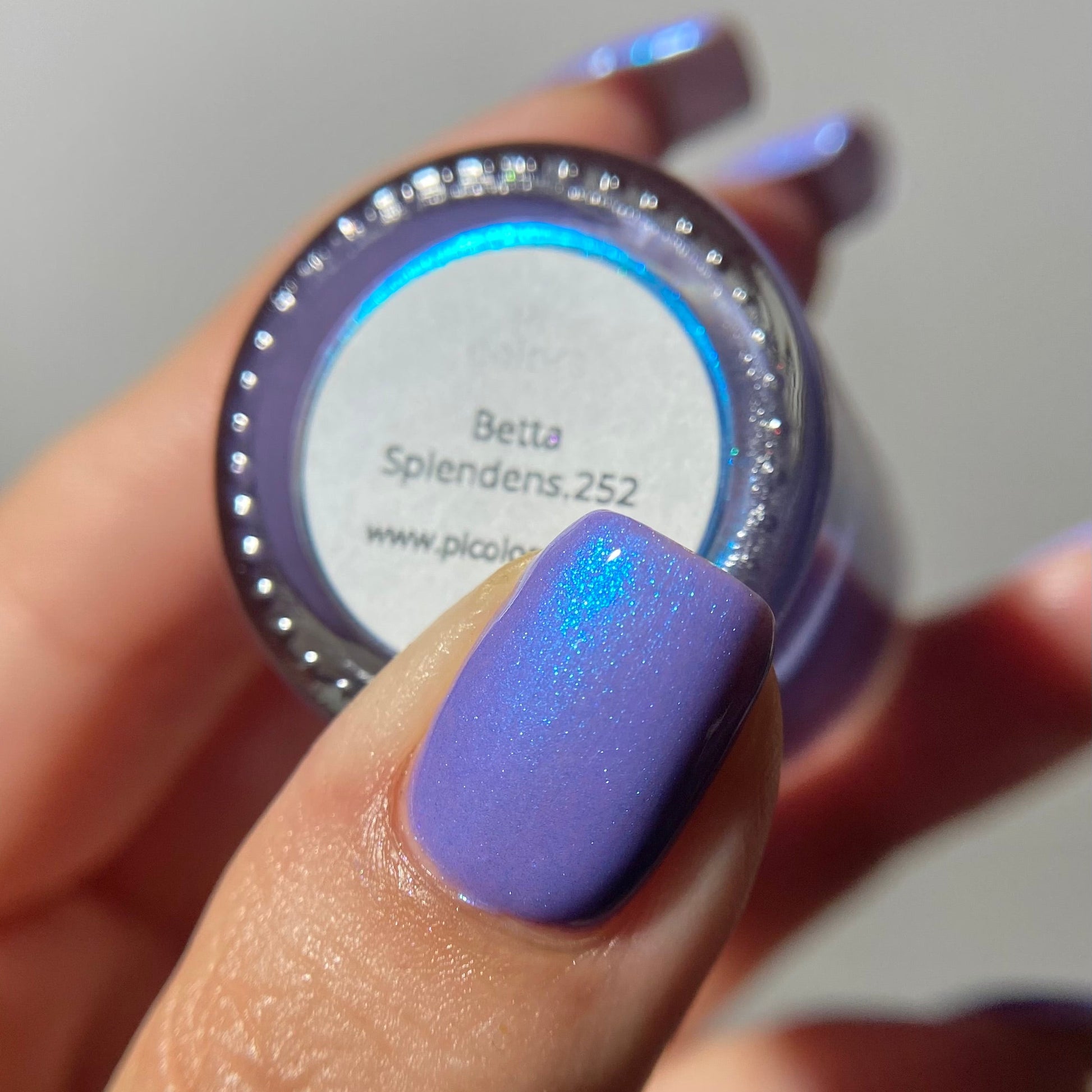 A close-up of a hand holding a small jar of nail polish or color swatch container, with the thumb showing a lavender-purple glittery nail finish. The label reads 'Betta Splendens.