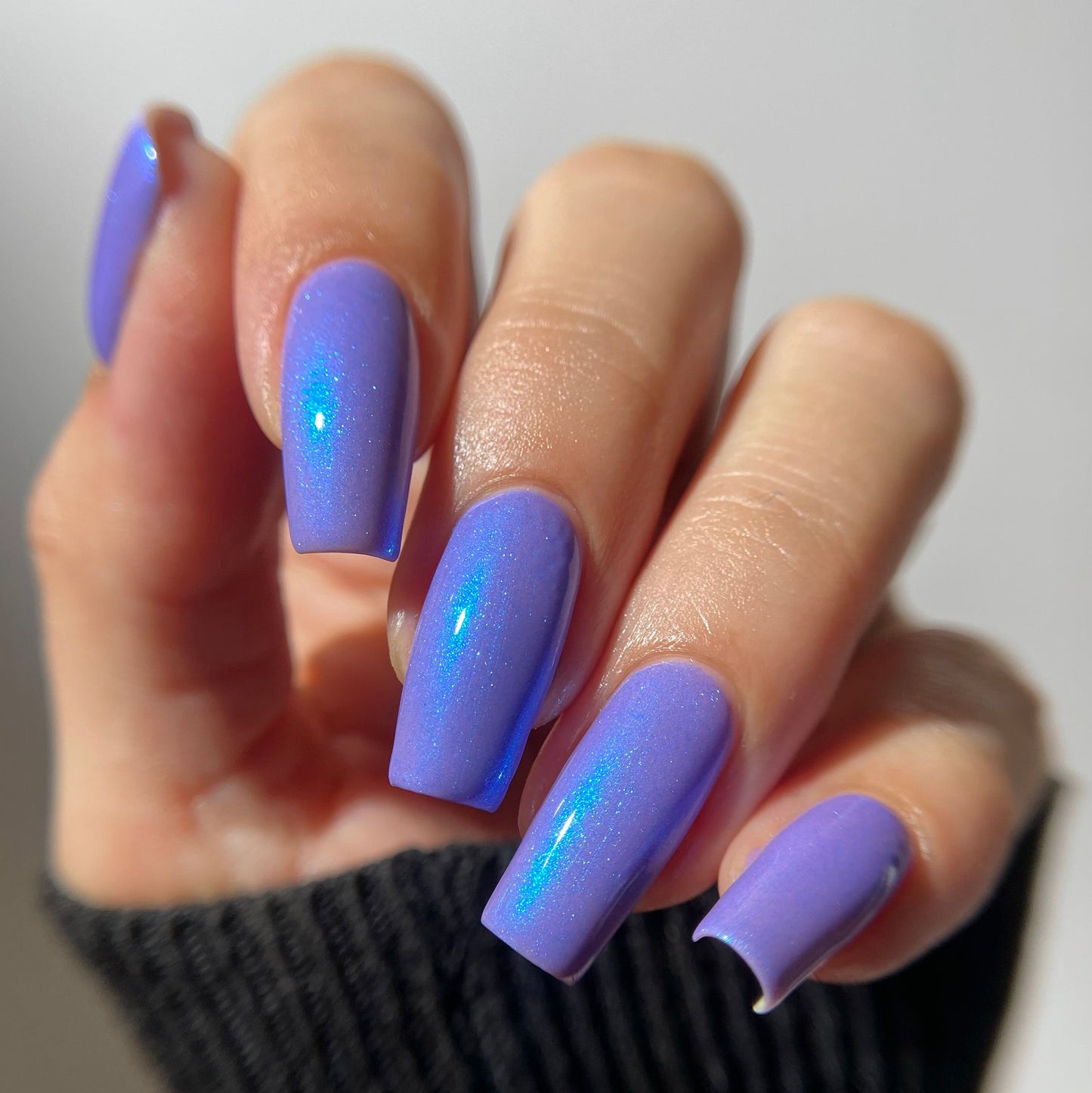 A close-up of a hand with long, almond-shaped nails painted in a light periwinkle purple color with a subtle iridescent shimmer, wearing a black ribbed knit sweater against a plain light background.