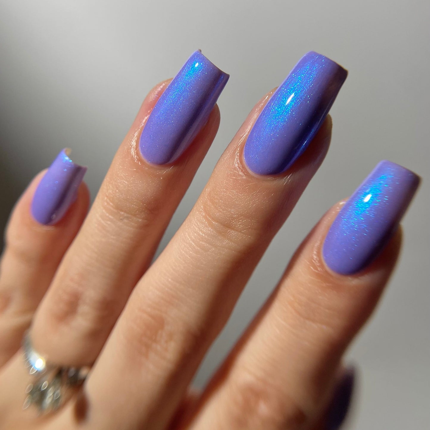 A close-up of a hand with long, square-shaped nails painted in a shimmering lavender-purple nail polish with a subtle iridescent blue cast.